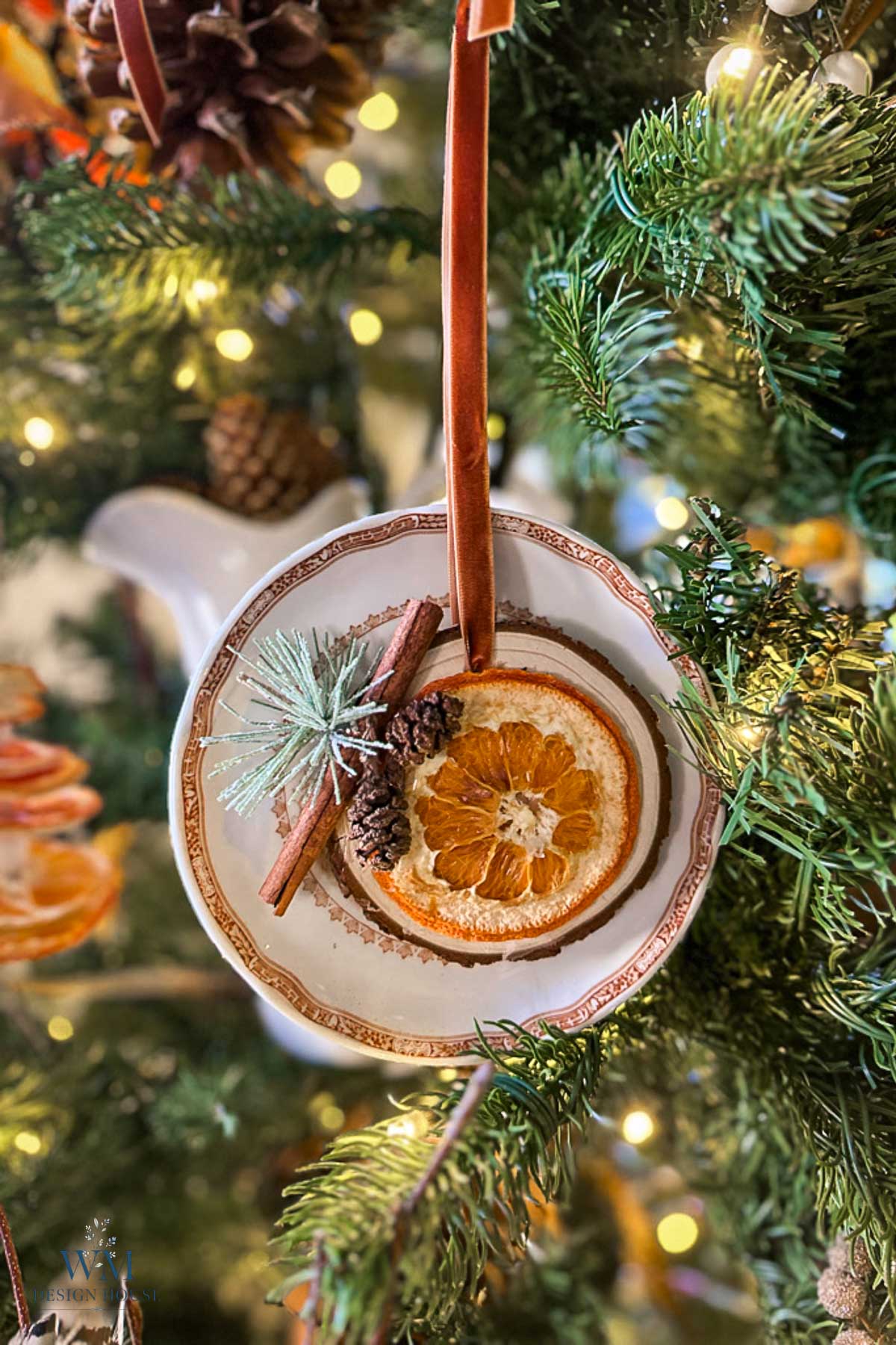 A Christmas tree ornament made from a small porcelain plate decorated with a dried orange slice, cinnamon stick, and mini pinecone, hanging by a velvet ribbon.