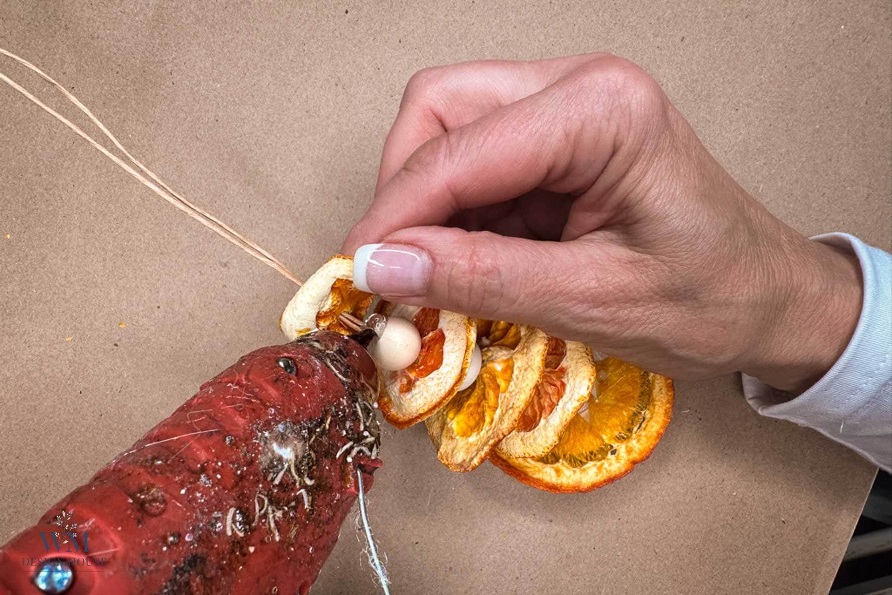 A hand uses a hot glue gun to attach a wooden bead between two dried orange slices.