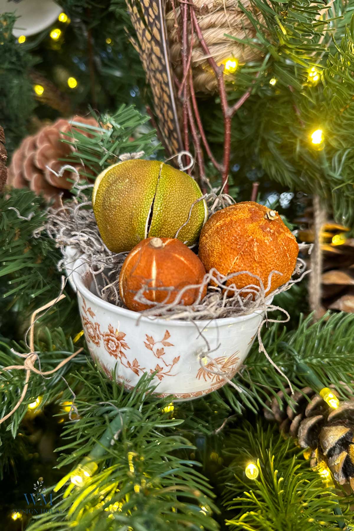 A small, patterned teacup filled with dried citrus fruits and nestling material, hanging as an ornament in a lit Christmas tree.