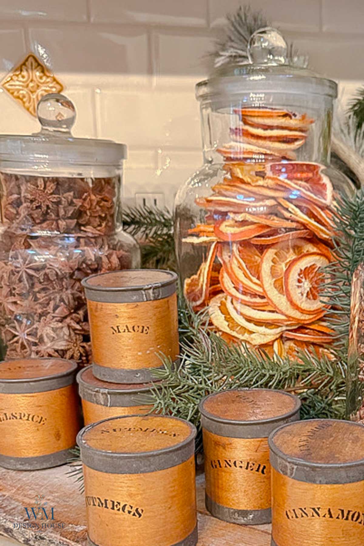 Glass jars filled with star anise and dried orange slices, surrounded by a group of labeled spice canisters including mace, allspice, nutmeg, ginger, and cinnamon.