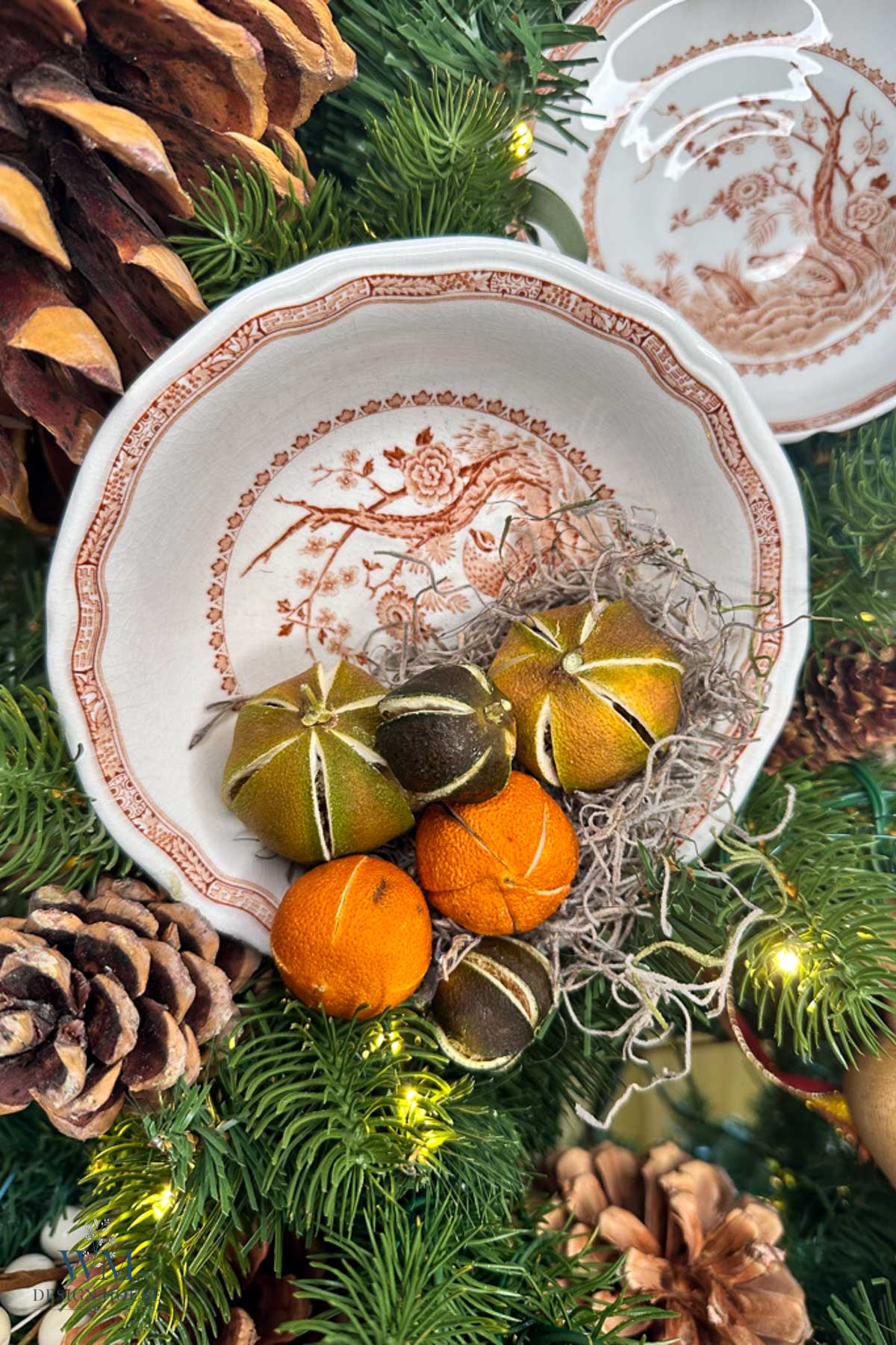 A shallow ceramic bowl with a brown floral pattern, filled with dried citrus fruits and Spanish moss, nestled among pine branches, pinecones, and string lights.