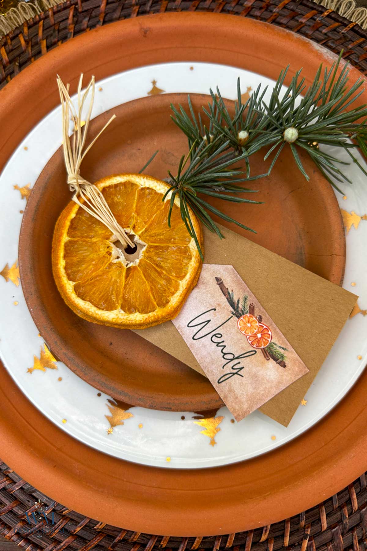 A place setting featuring a terracotta plate, a dried orange slice, a pine sprig, and a place card labeled “Wendy.”
