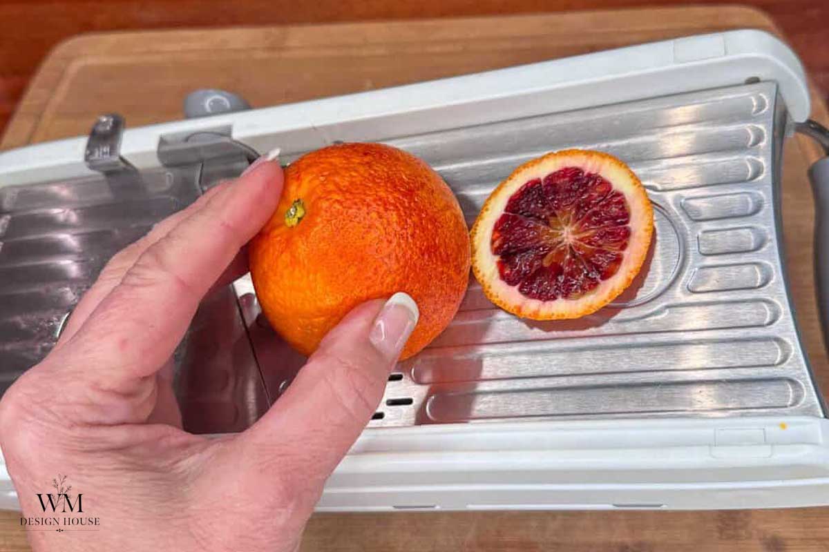 A hand holds a whole blood orange next to a sliced half on a metal mandoline slicer, preparing the fruit to make dehydrated orange slices.