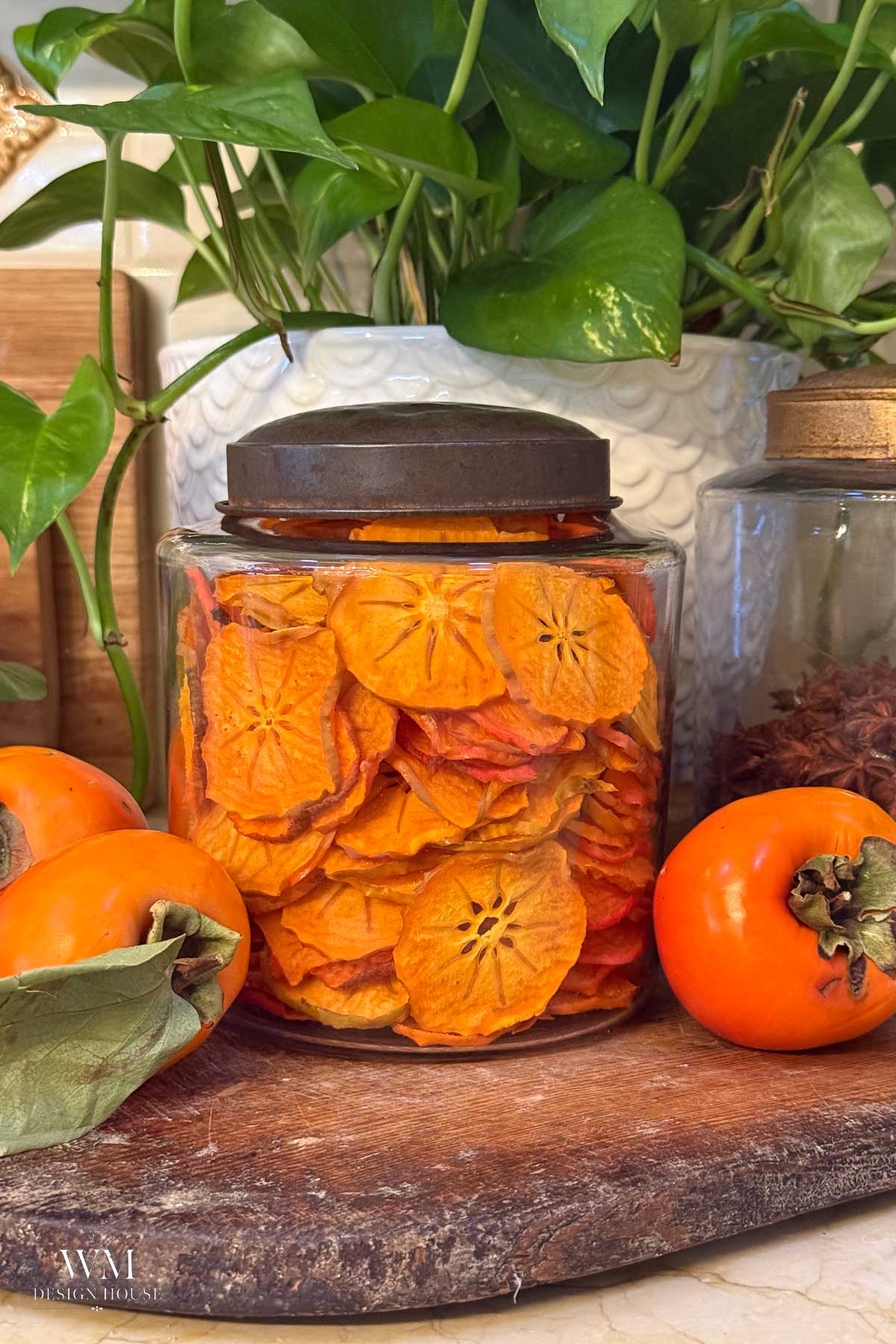 A glass jar filled with dried persimmon slices, flanked by two fresh whole persimmons on a wooden surface.