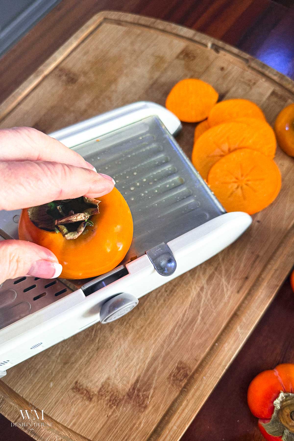 A hand using a mandoline to slice a bright orange persimmon on a wooden cutting board, with several already sliced pieces nearby.