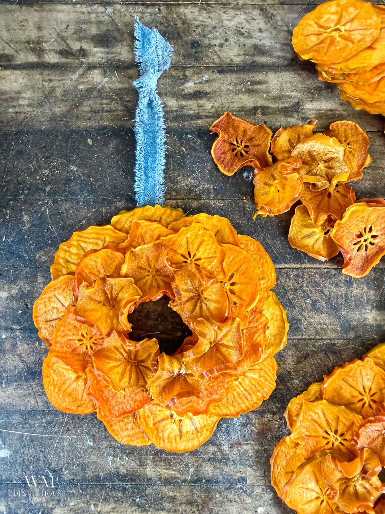 A wreath made of dried, sliced persimmons hanging from a blue frayed fabric ribbon, surrounded by loose slices on a rustic wooden surface.