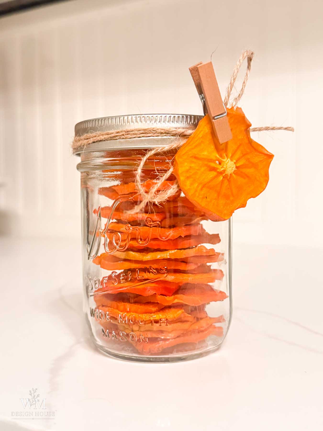 A glass Mason jar filled with dried persimmon slices, tied with twine and a wooden clothespin holding a single slice to the lid.