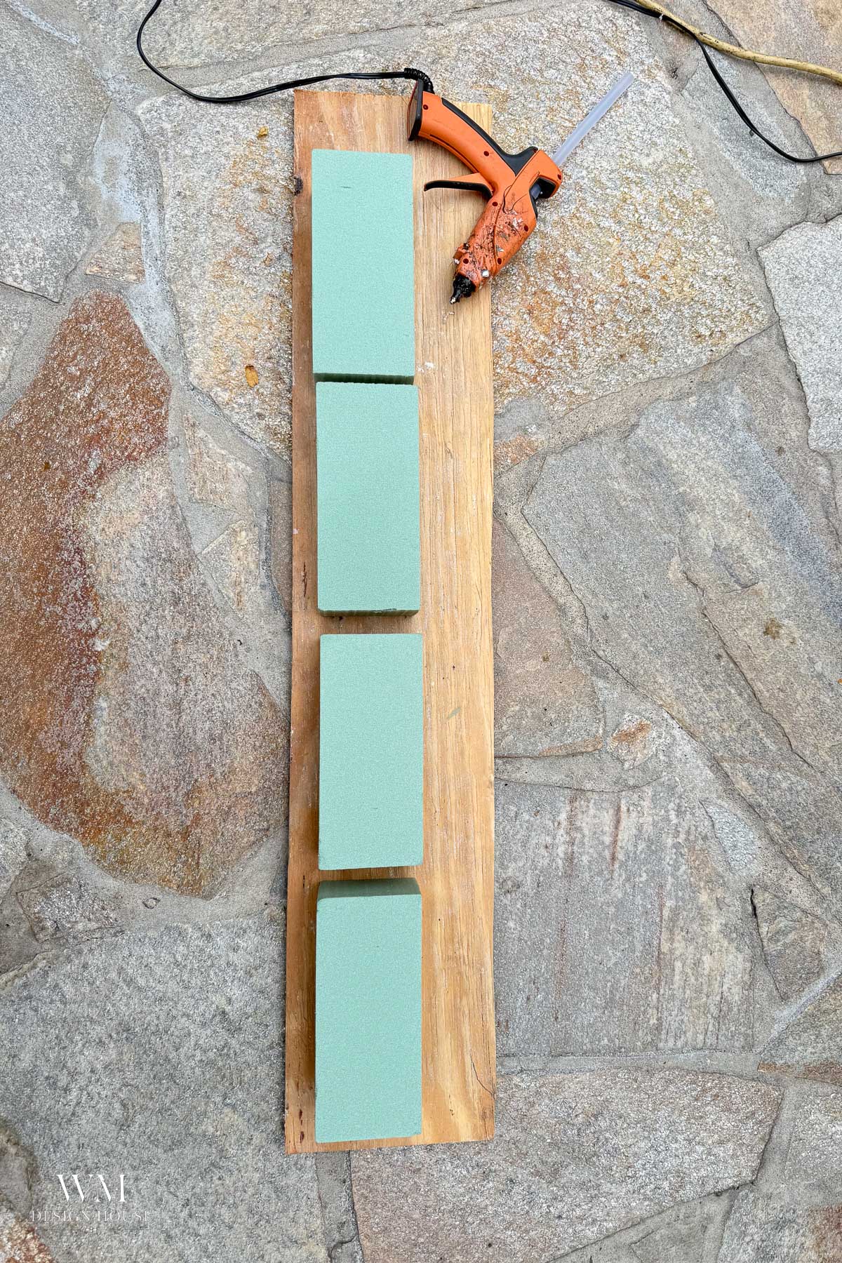 Four rectangular green floral foam blocks arranged in a line on a wooden board for fall mantel decor, with a glue gun resting nearby.