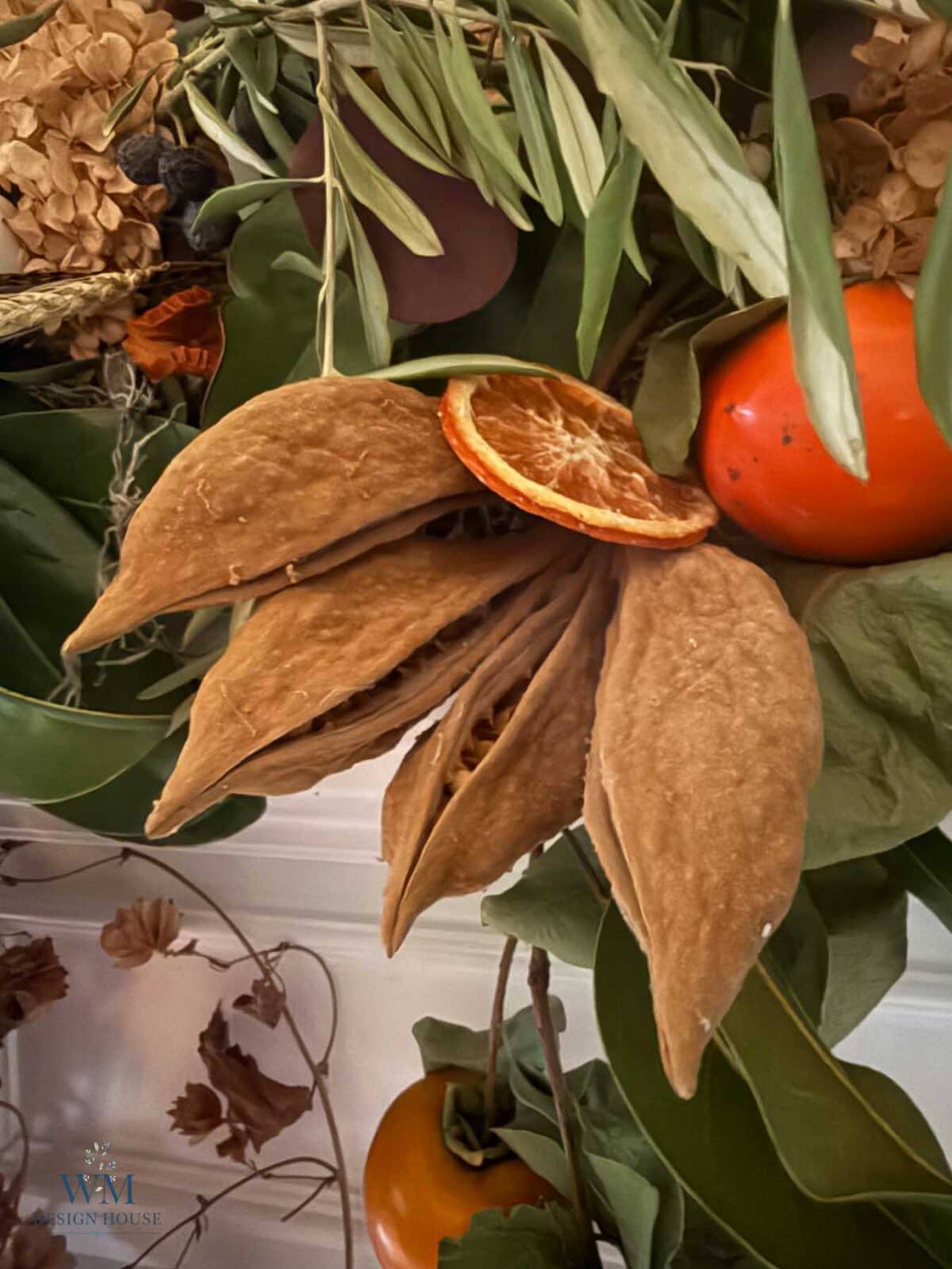 A seasonal fall mantel arrangement featuring large brown seed pods, orange slices, and persimmons among green foliage.