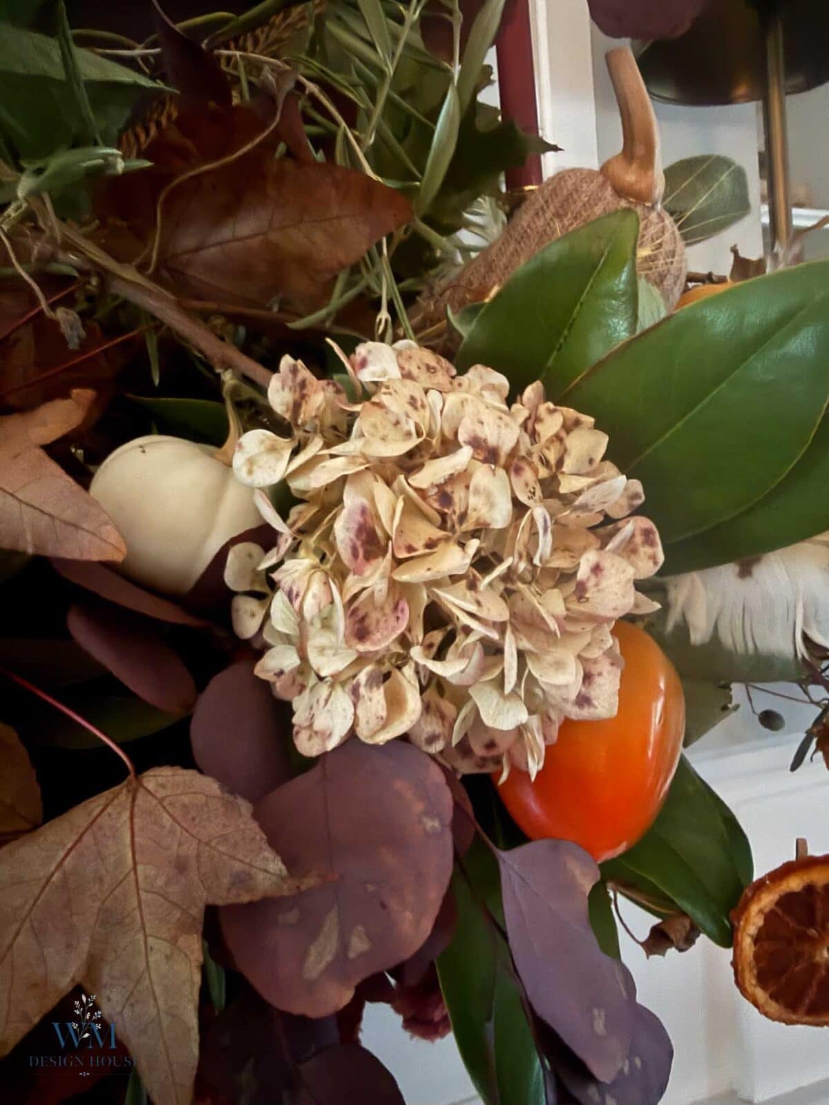 A fall mantel arrangement featuring a dried hydrangea blossom, green leaves, small pumpkins, and dried fruit slices for seasonal home styling.