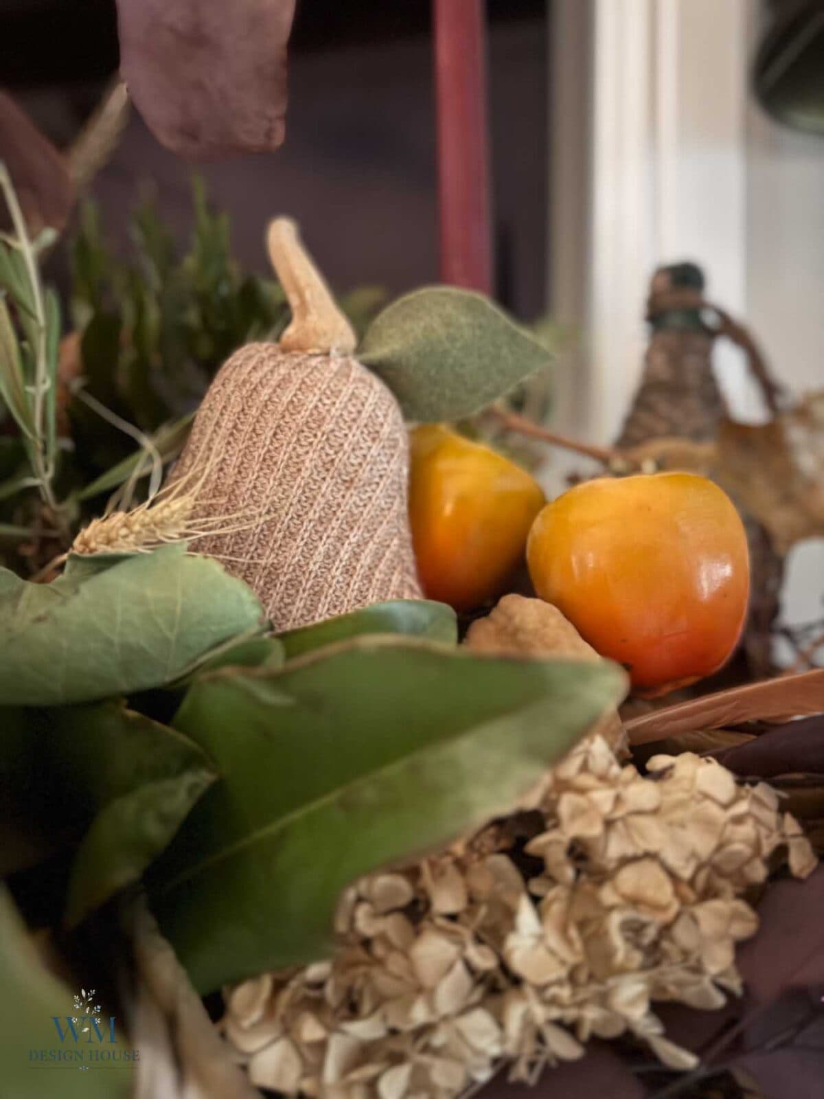 A textured fabric pumpkin ornament nestled among greenery, dried flowers, and two persimmons for fall mantel decor.