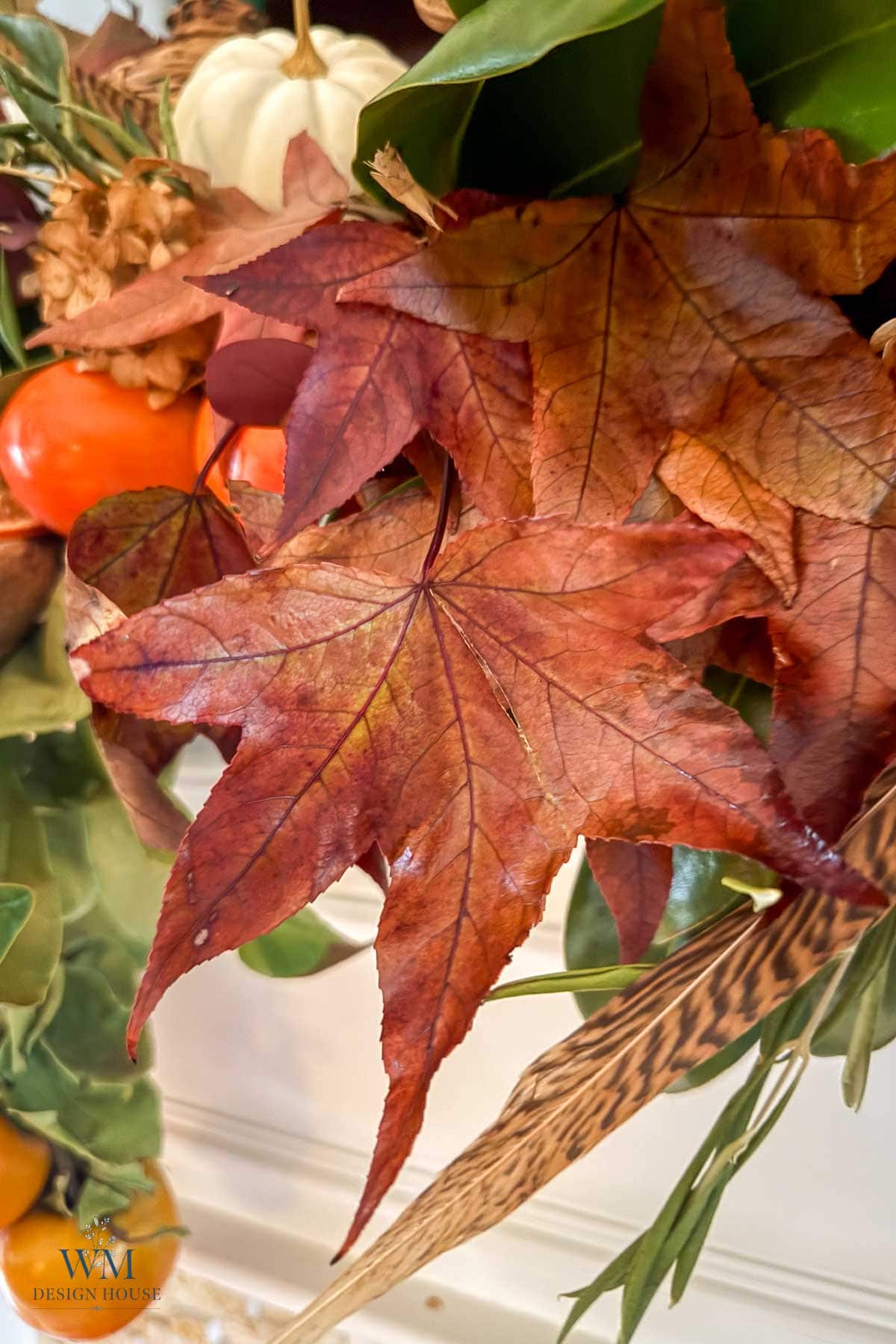 A close-up of a fall home decor wreath featuring red maple leaves, orange fruits, and a small white pumpkin, with a WM Design House logo in the bottom corner.