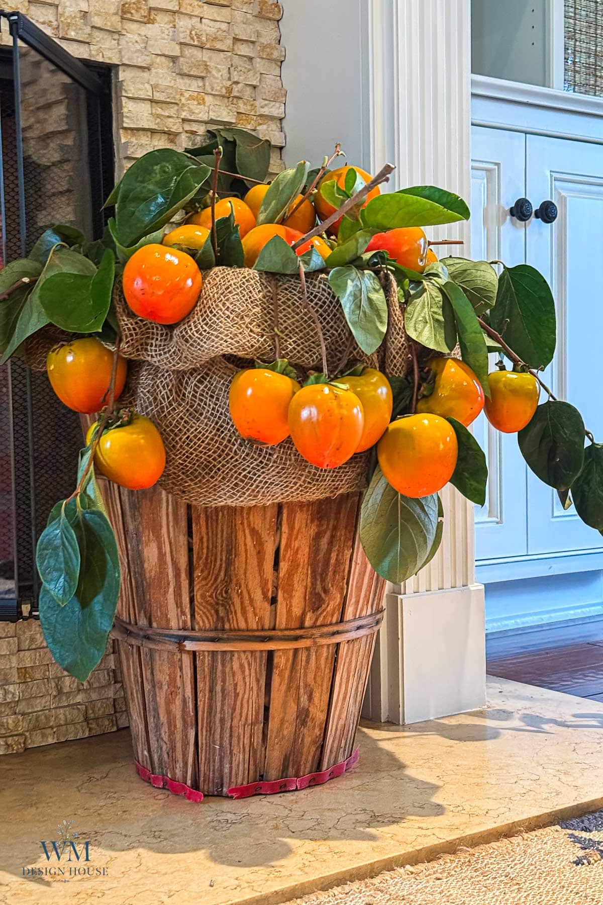 A wooden basket filled with branches of ripe orange persimmons and green leaves, partially wrapped in burlap for fall mantel decor.