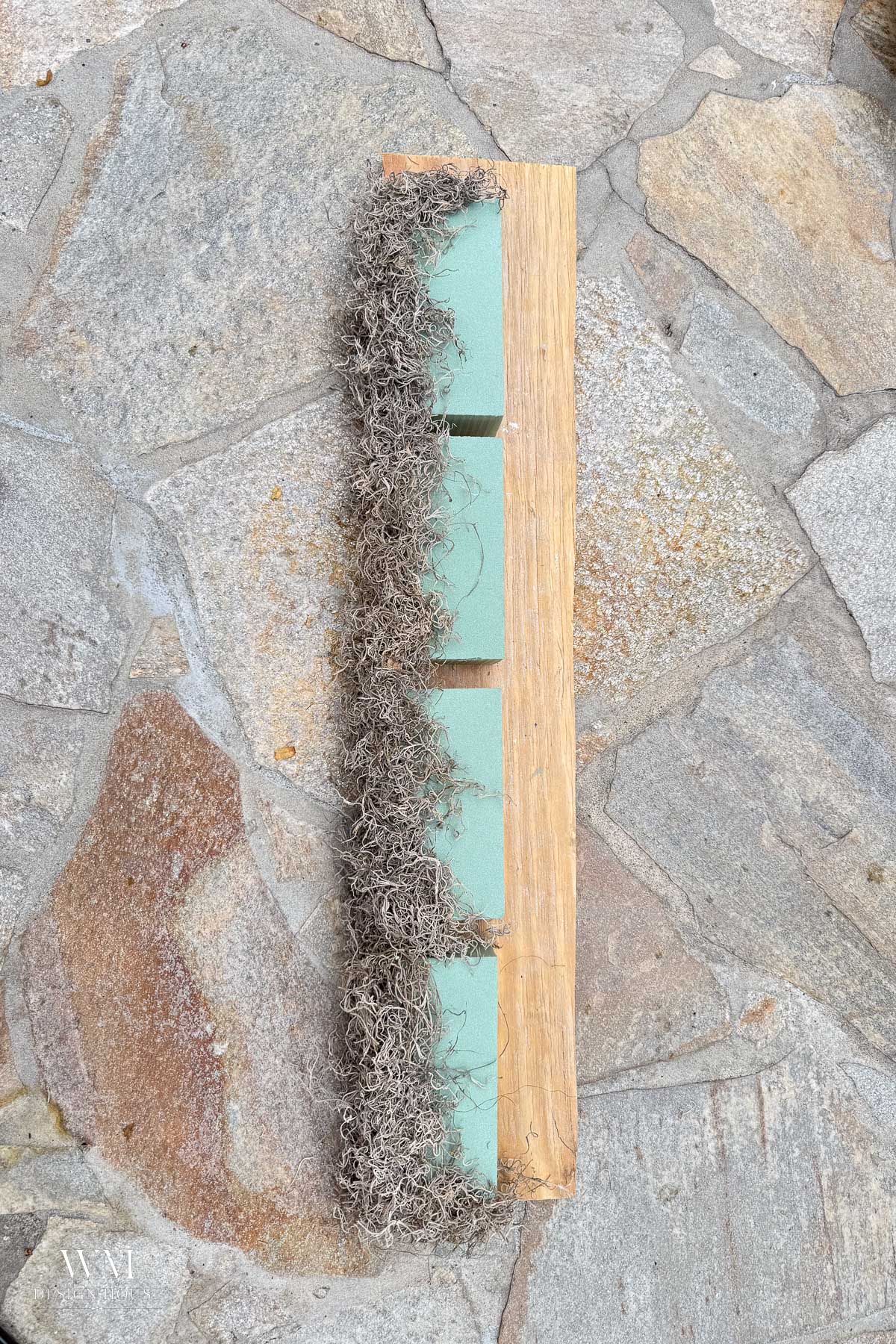 A row of green floral foam bricks decorated with dried Spanish moss for fall home styling, attached to a narrow wooden board on a stone patio.