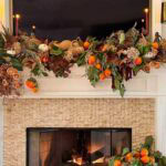 A fall mantel decorated with a garland of dried leaves, oranges, pumpkins, and candles for seasonal home decor.