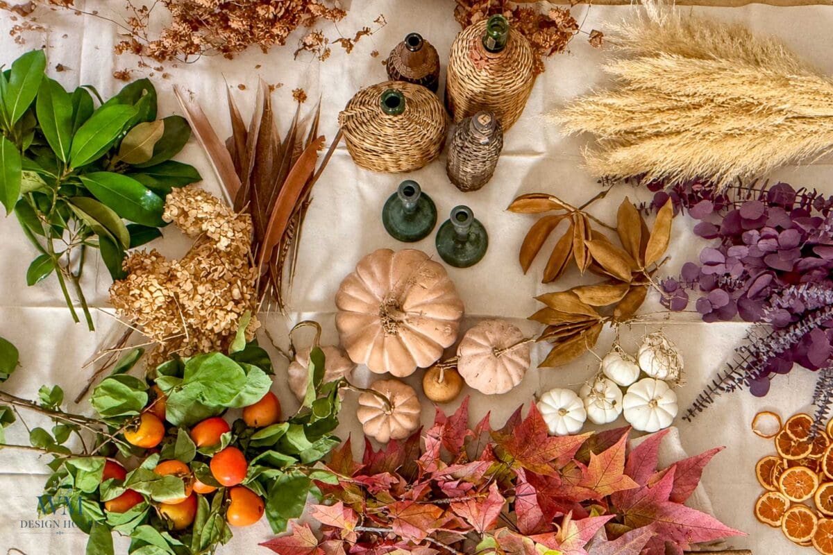 An overhead view of DIY mantel decor supplies, including decorative pumpkins, persimmons, dried grasses, woven vases, and fallen leaves spread across a light-colored surface for foraged fall decor.