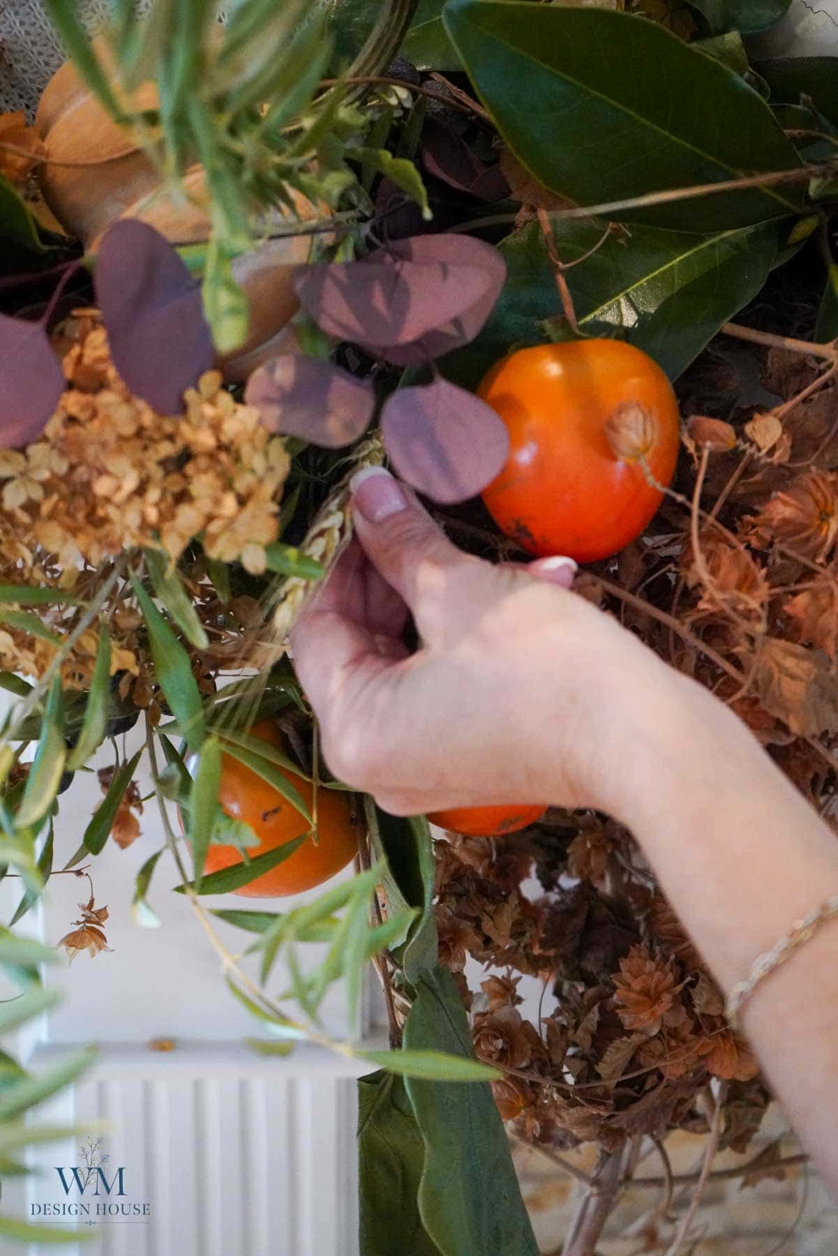 A hand adjusts an arrangement of dried flowers, green leaves, and bright orange persimmons as part of a fall home decor display.