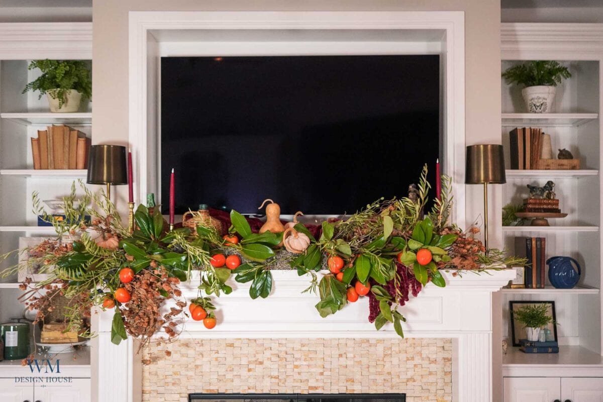 A fall mantel decorated with a lush garland of greenery, persimmons, dried branches, and a gourd, positioned beneath a mounted television.