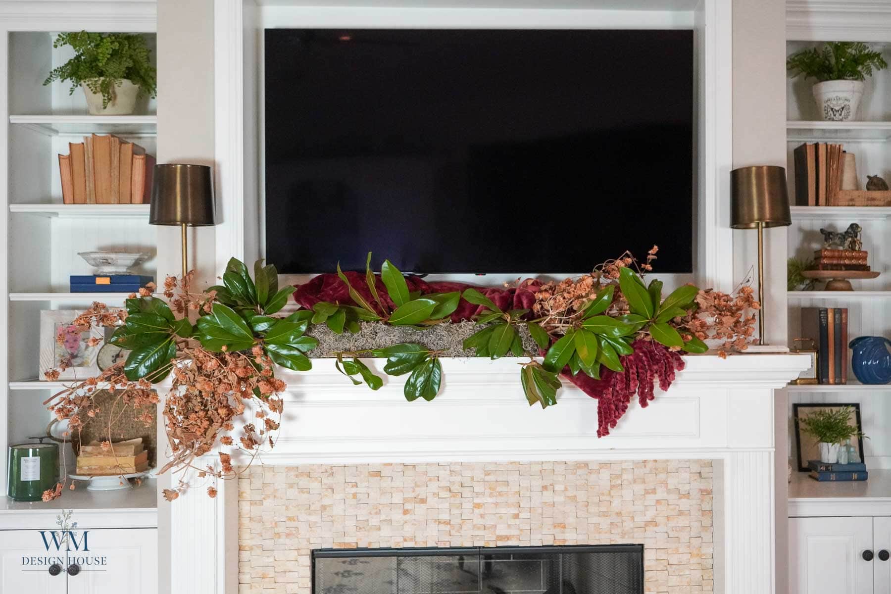 A fall mantel decorated with a garland of magnolia leaves, dried flowers, and burgundy velvet fabric, situated beneath a wall-mounted television and framed by built-in shelves for seasonal decorating.