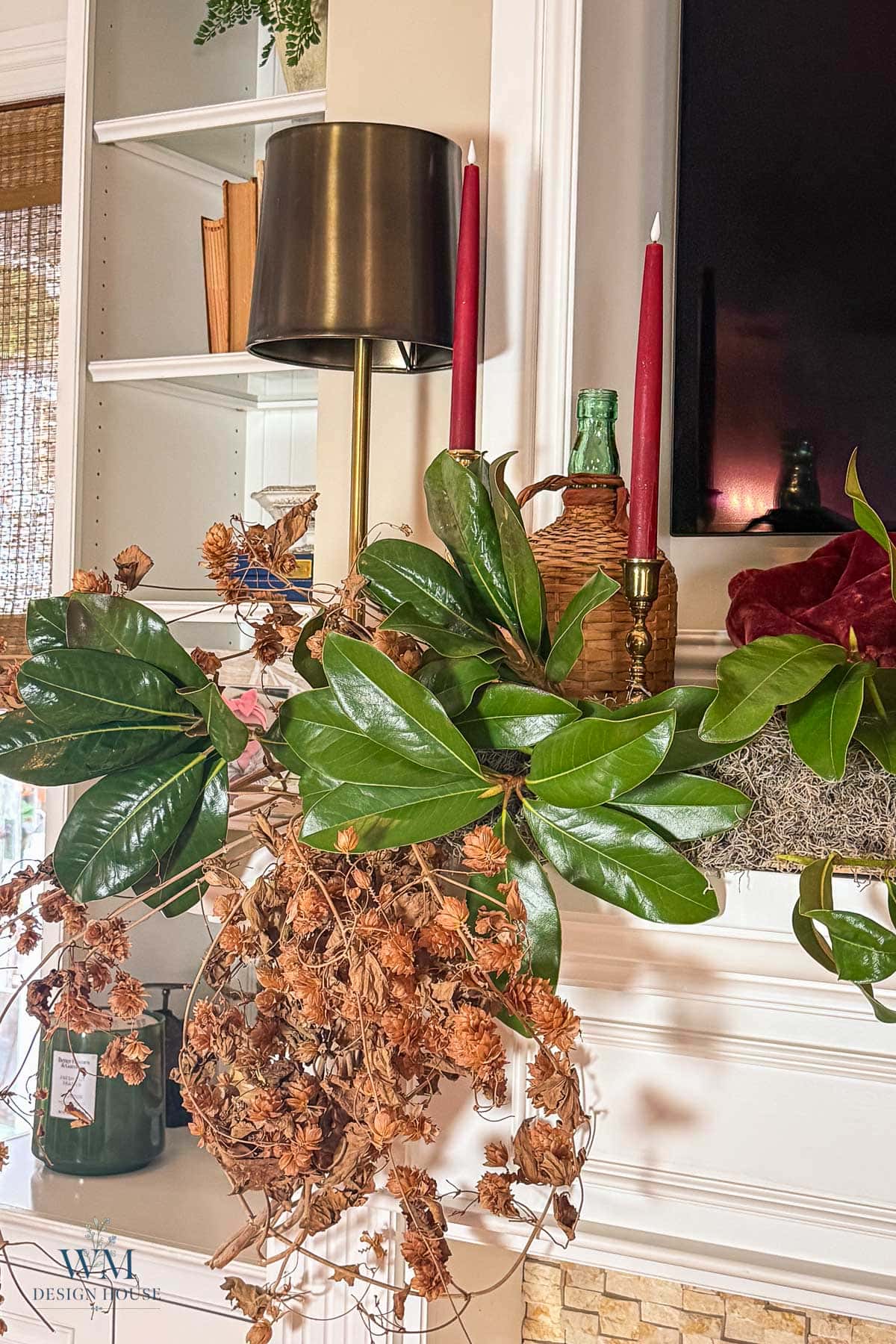A fall mantel styled with magnolia branches, dried hops, two red taper candles in brass holders, and a glass lamp.