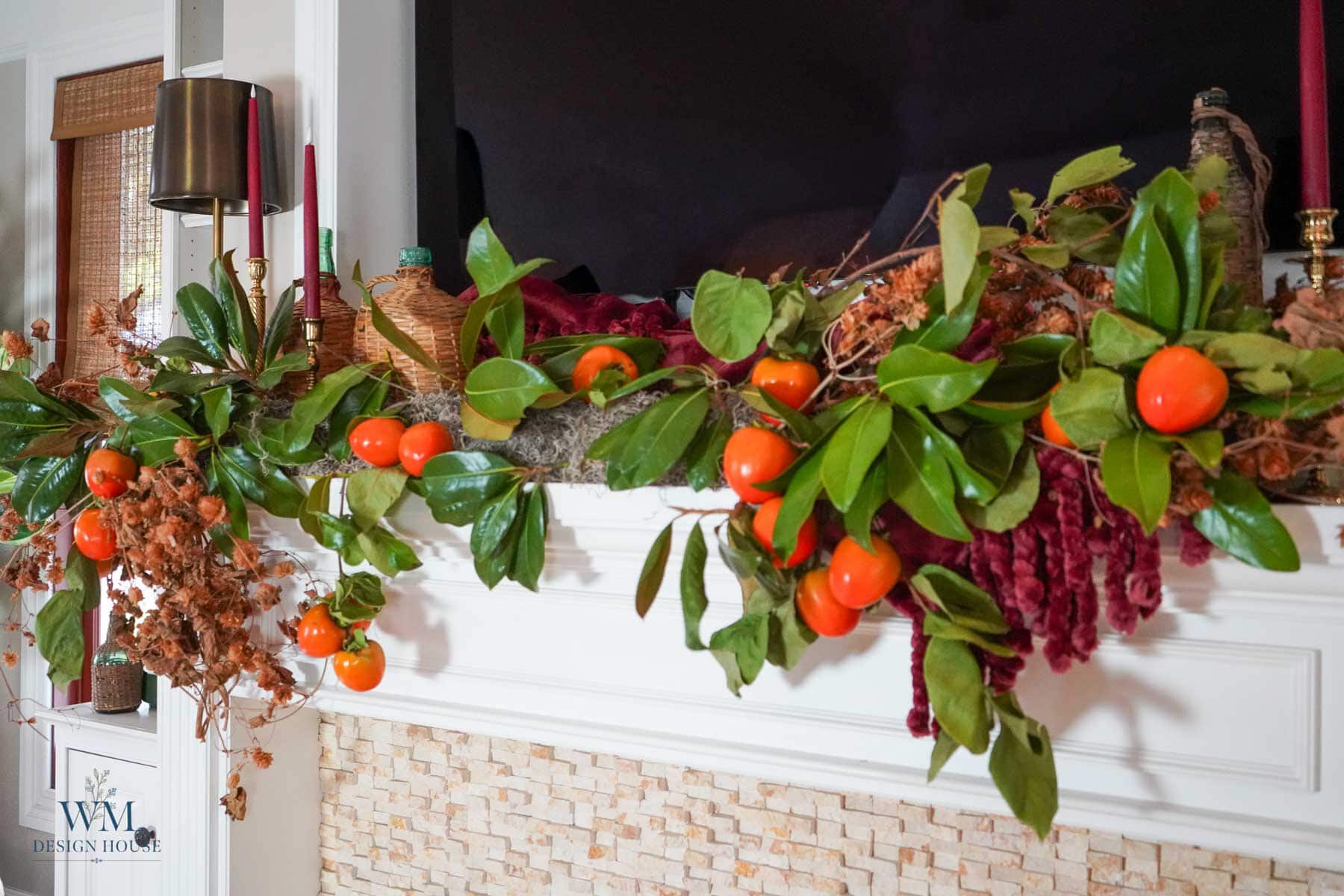 A white fireplace mantel styled with fall decor featuring a garland of persimmons, green leaves, and dried flowers.