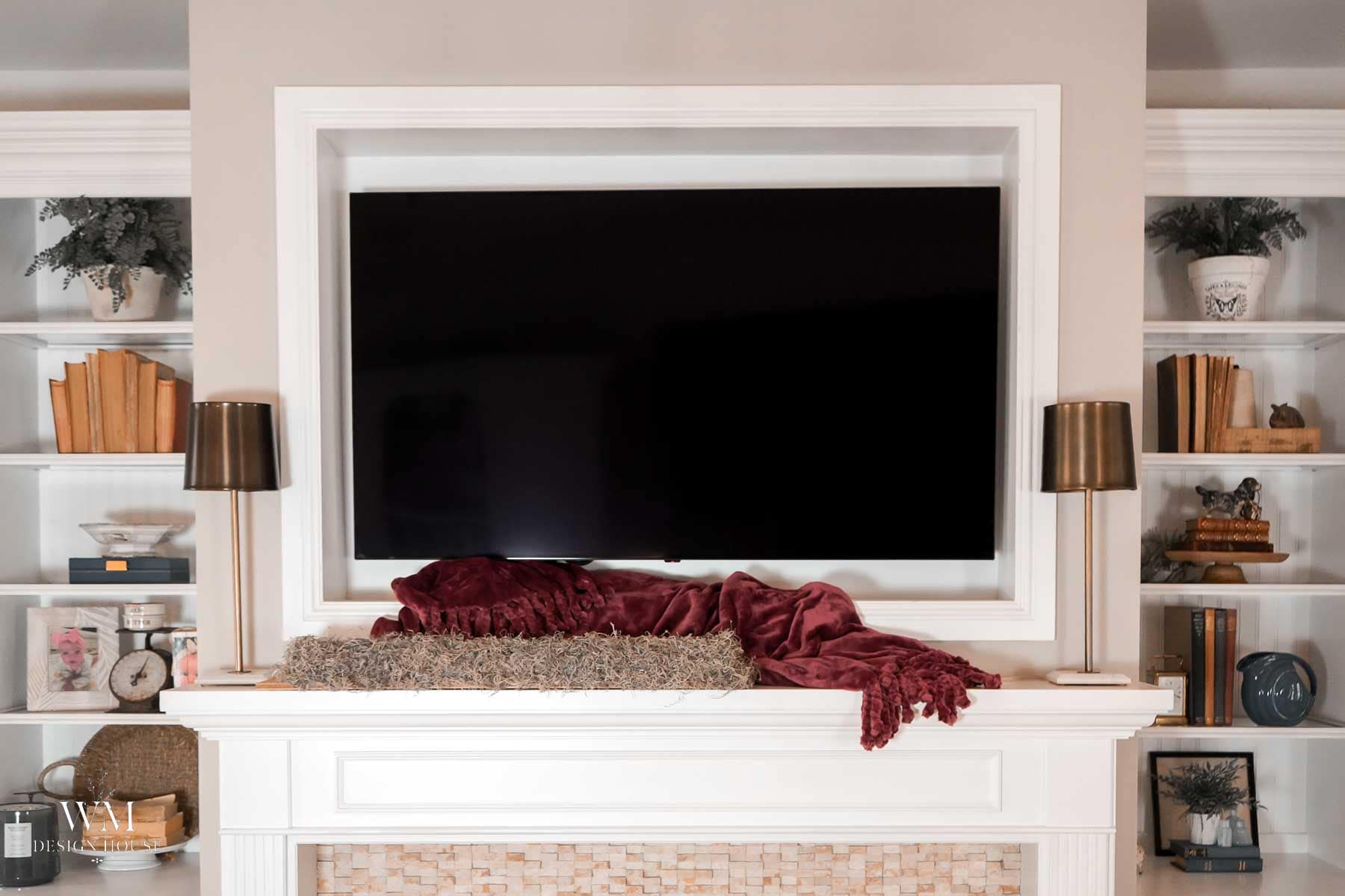 A flat-screen television mounted above a white fireplace mantel styled with fall decor, featuring a maroon throw blanket, two gold lamps, and flanking shelves filled with books and accents.