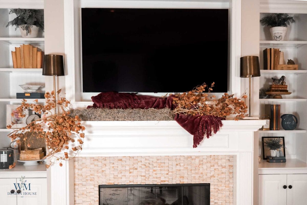 A fireplace mantel styled with fall decor, featuring dried hops, a deep red velvet throw, and two lamps flanking a wall-mounted television between built-in bookshelves.