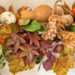 A decorative arrangement featuring dried leaves, assorted gourds, small orange fruits, dried citrus slices, and sprigs of greenery.