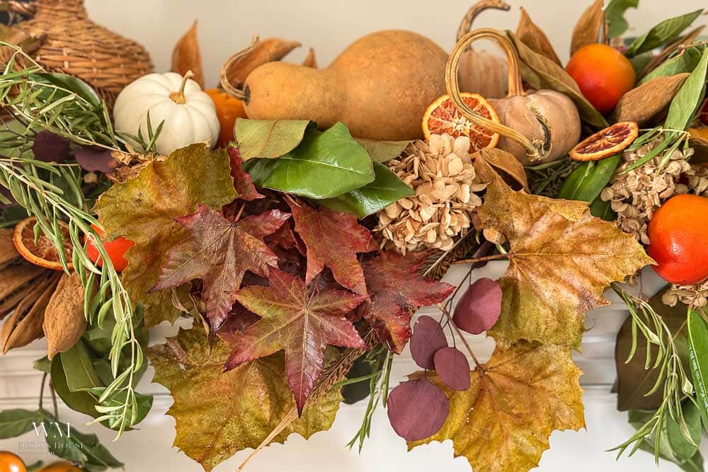 A decorative arrangement featuring dried leaves, assorted gourds, small orange fruits, dried citrus slices, and sprigs of greenery.