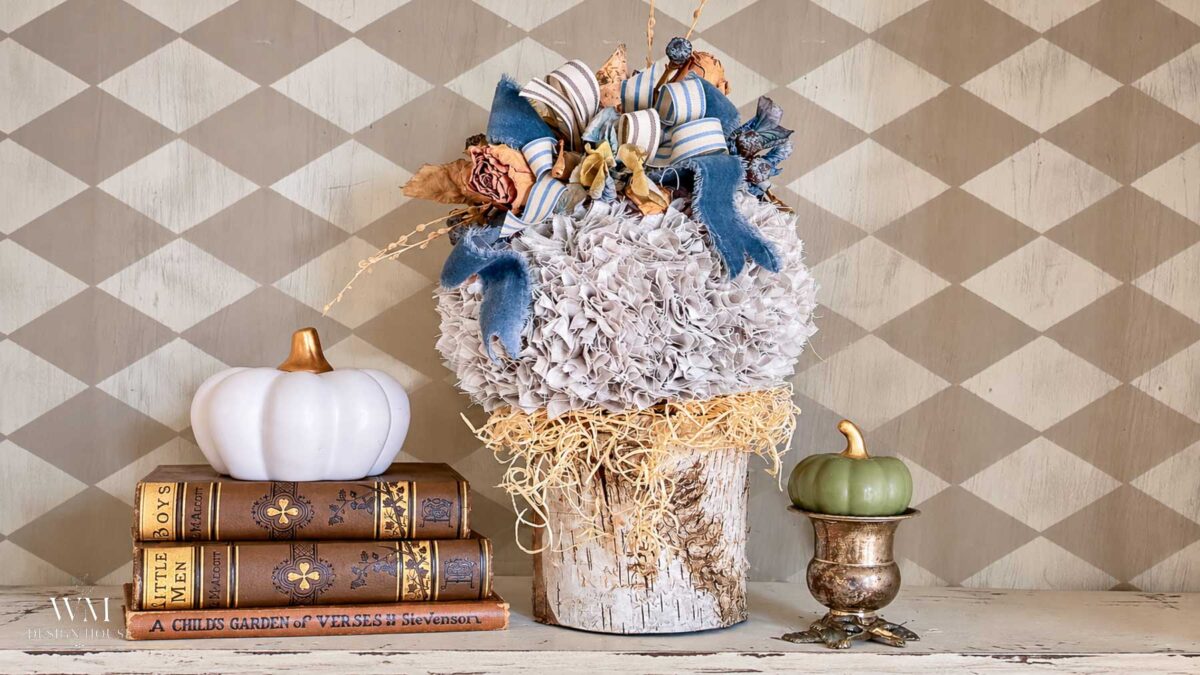 A decorative arrangement featuring a no sew fabric pumpkin topped with blue and tan ribbons on a birch log, flanked by a white pumpkin, vintage books, and a green pumpkin in a metallic pedestal bowl.