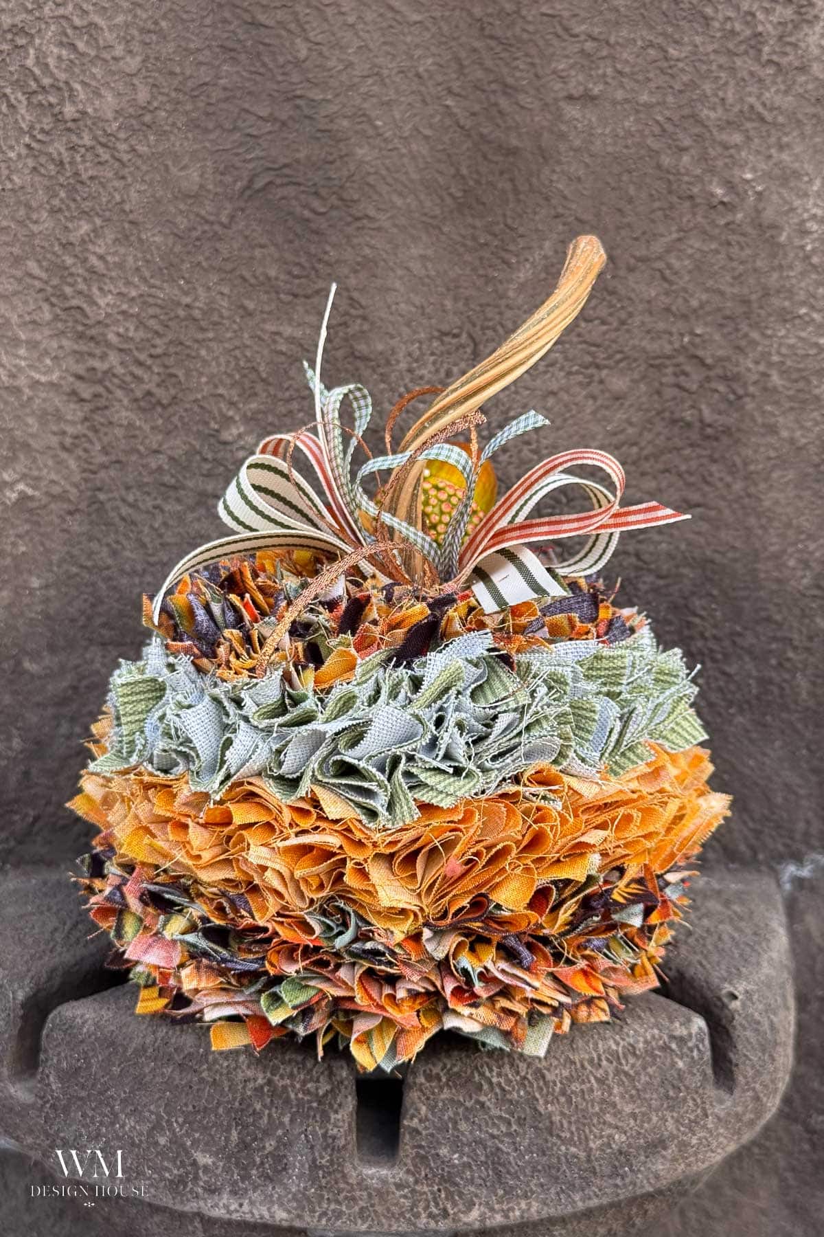 No sew fabric pumpkin decoration made of layered, ruffled squares of fabric in shades of orange, green, and brown, topped with a decorative striped bow and a dried pumpkin stem.