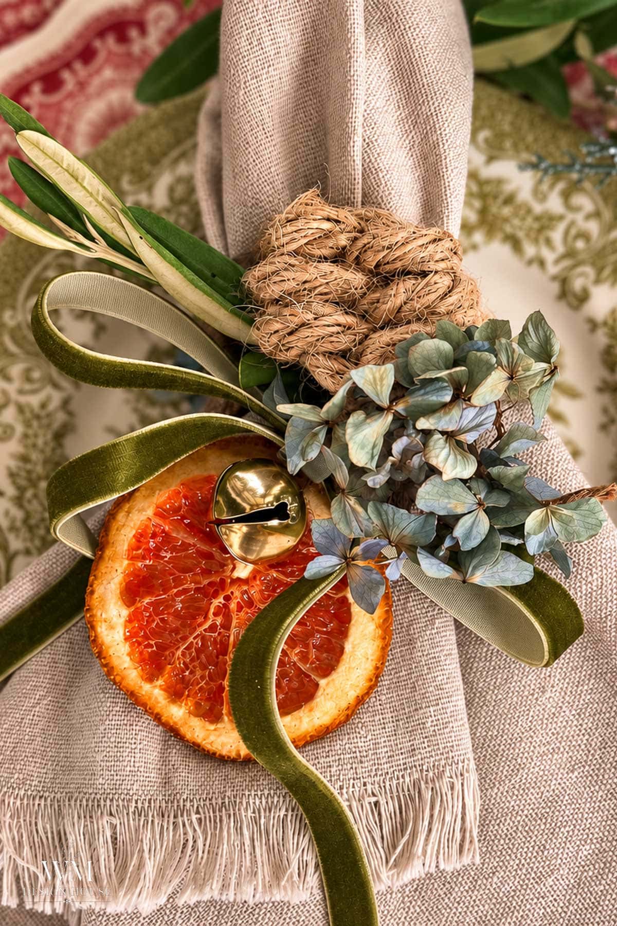 A linen napkin tied with a green velvet ribbon and a decorative leaf knot, featuring a dried orange slice, a brass bell, and a small sprig of blue hydrangea in a set of Christmas napkin rings.