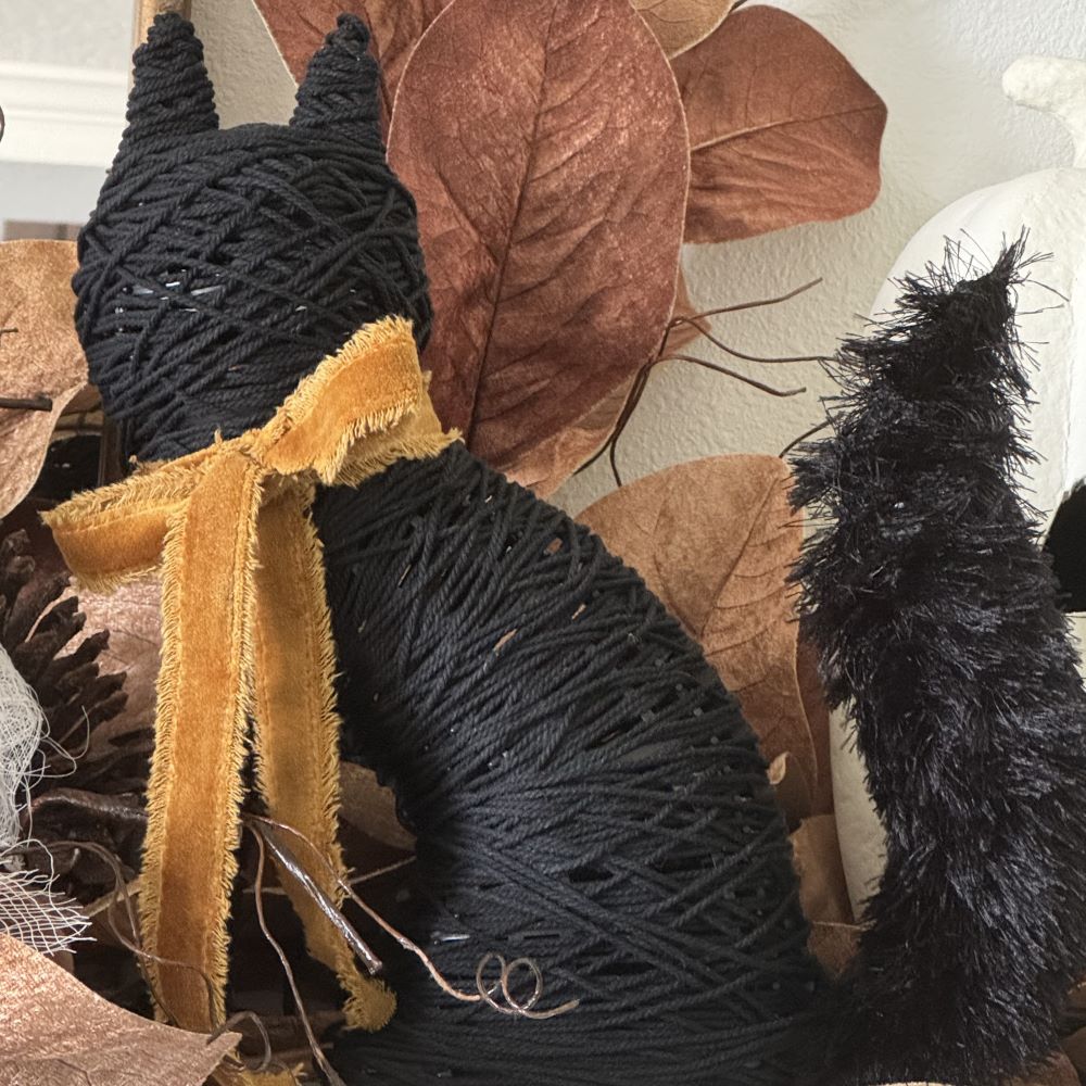 A decorative black yarn-wrapped cat figure with a gold ribbon tied around its neck, set against a background of brown faux leaves.