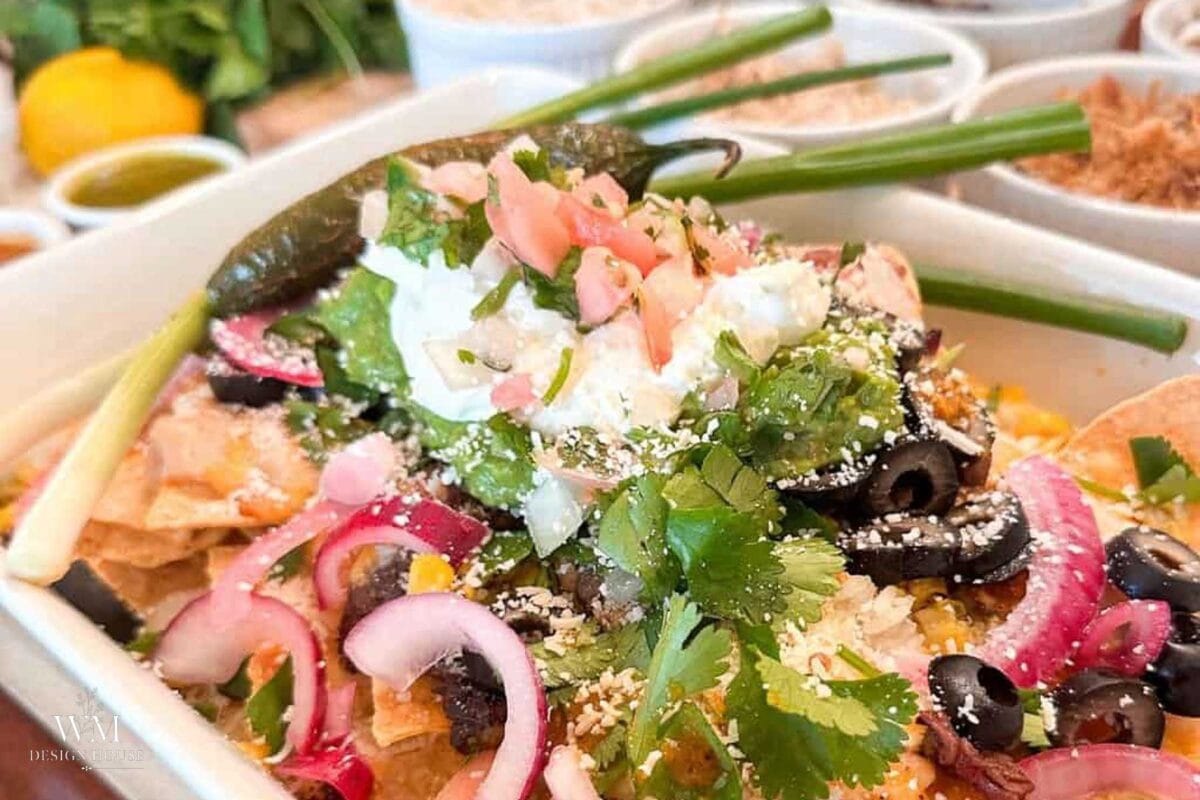 A bowl of homemade nachos topped with guacamole, sour cream, black olives, pickled red onions, cilantro, and a roasted jalapeño.