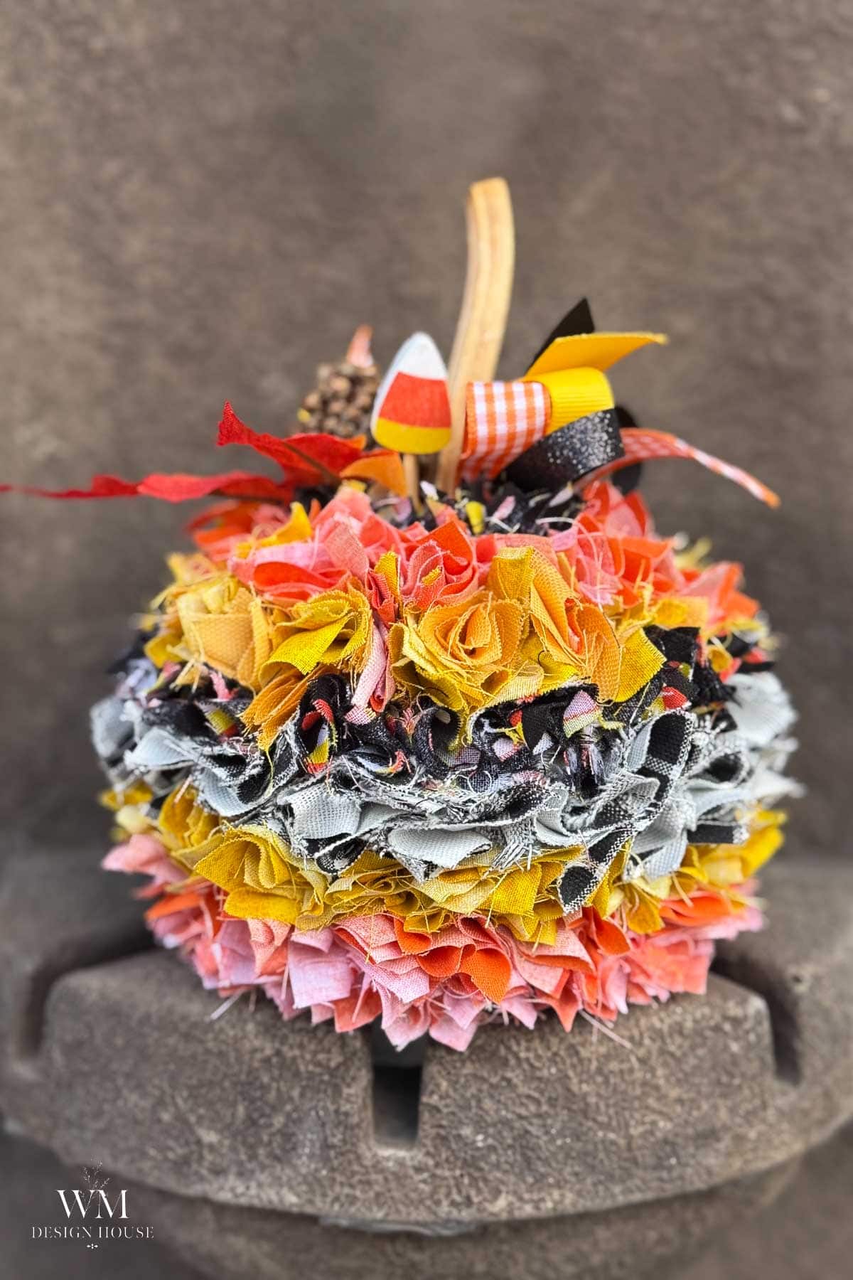 A handmade fabric pumpkin decoration layered in orange, yellow, black, and white strips, topped with a natural stem, a candy corn cutout, and ribbons.
