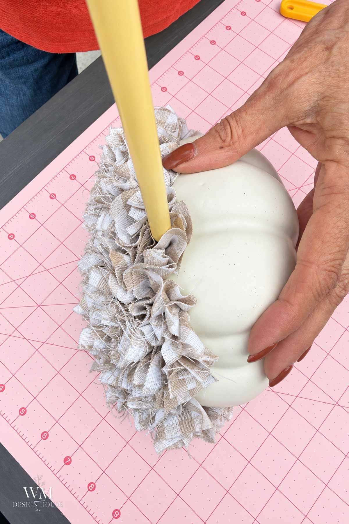 A beige kitting needle tool pushes a piece of tan and white gingham fabric into a hole into a foam pumpkin.
