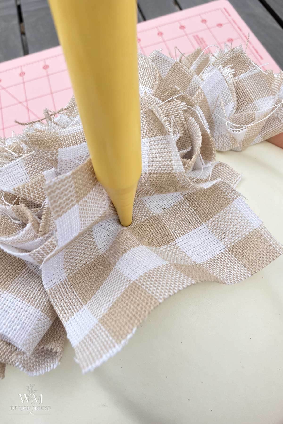 A beige kitting needle tool pushes a piece of tan and white gingham fabric into a hole into a foam pumpkin.