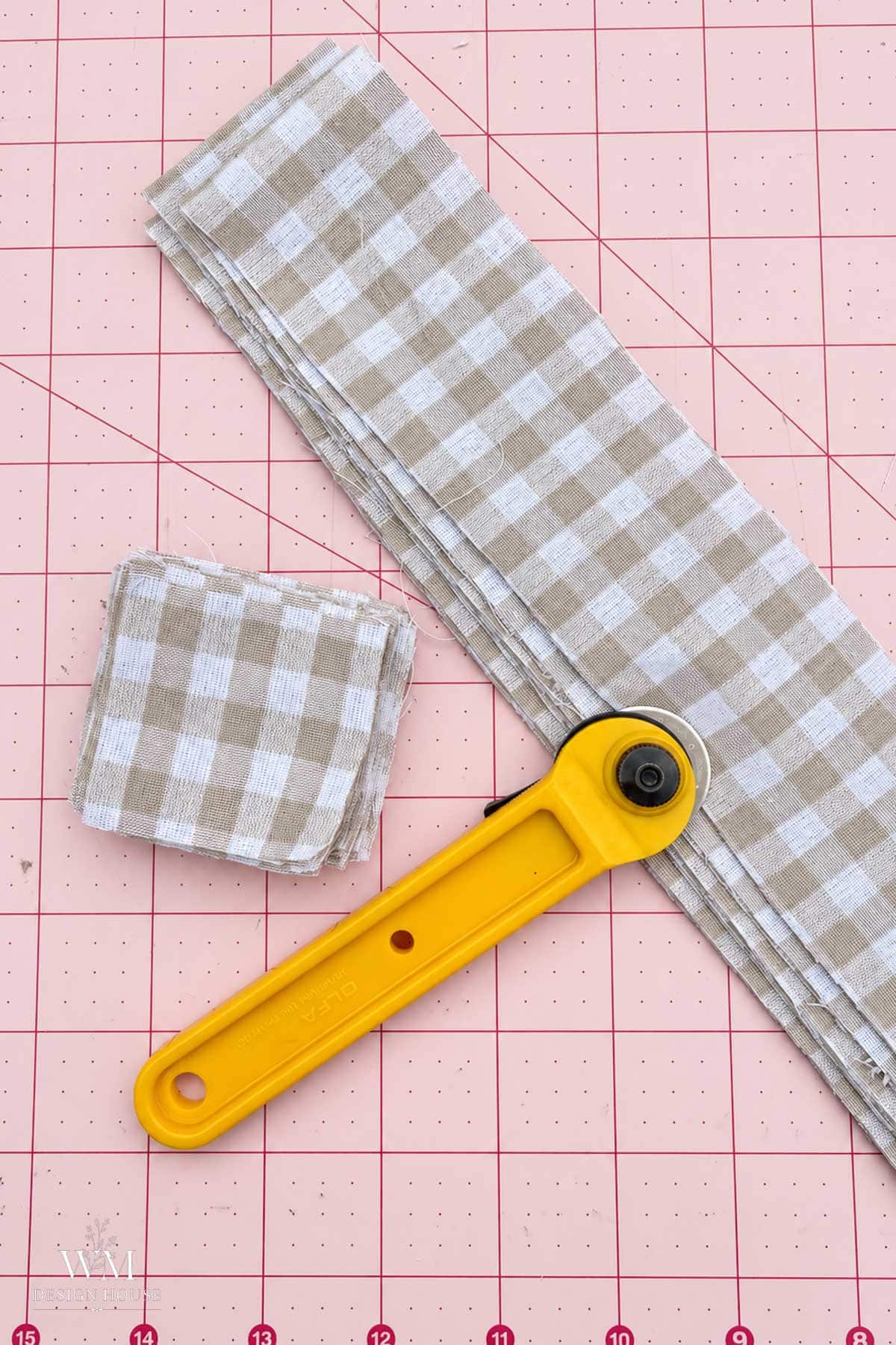 A yellow rotary cutter on a pink cutting mat next to a stack of fabric strips and a small stack of squares, all in a beige and white gingham pattern to make a no sew fabric pumpkin.