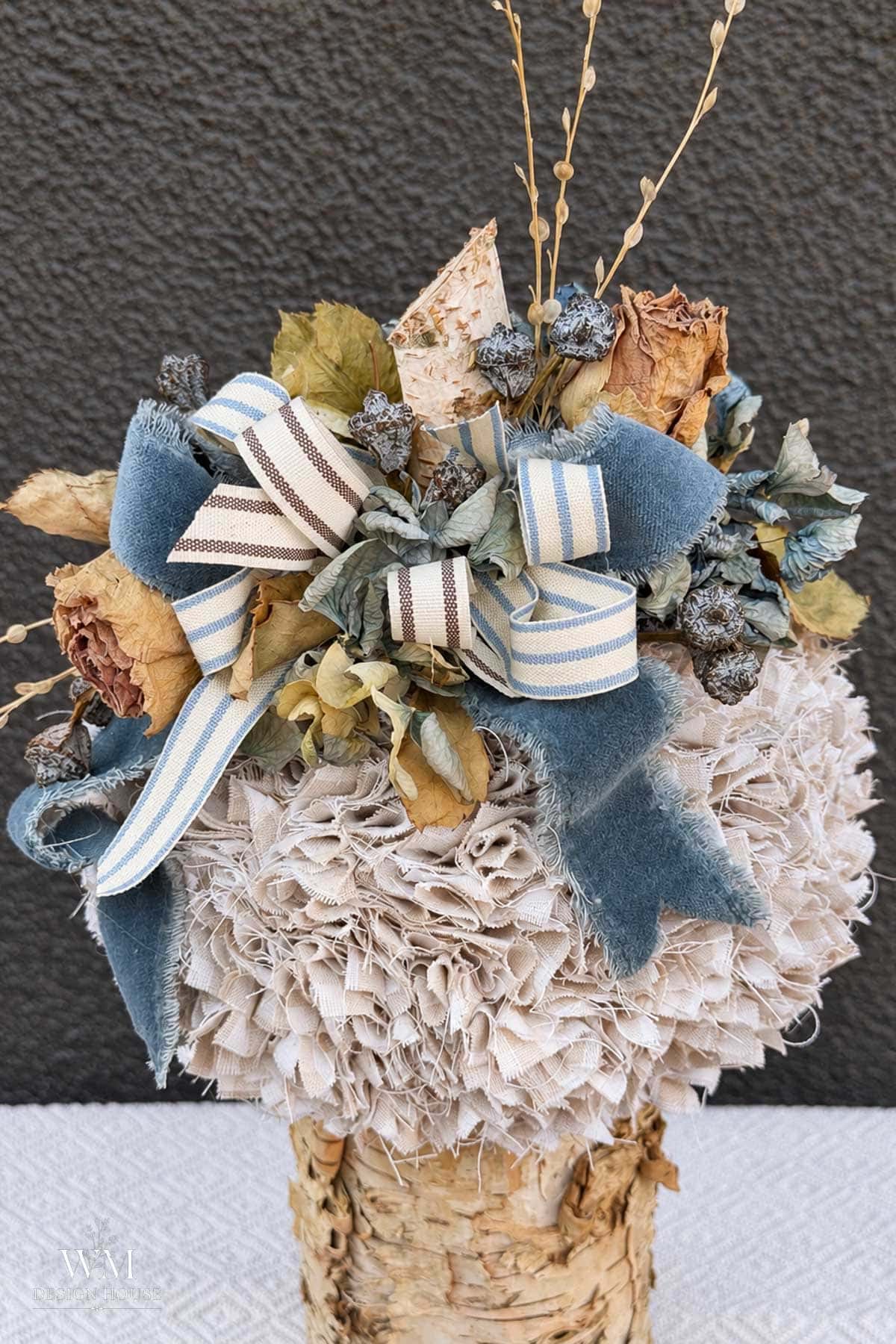 A decorative fabric no sew pumpkin made of 2" fabric squares, topped with dried roses, greenery, and blue and striped ribbons, perched on a birch bark base.