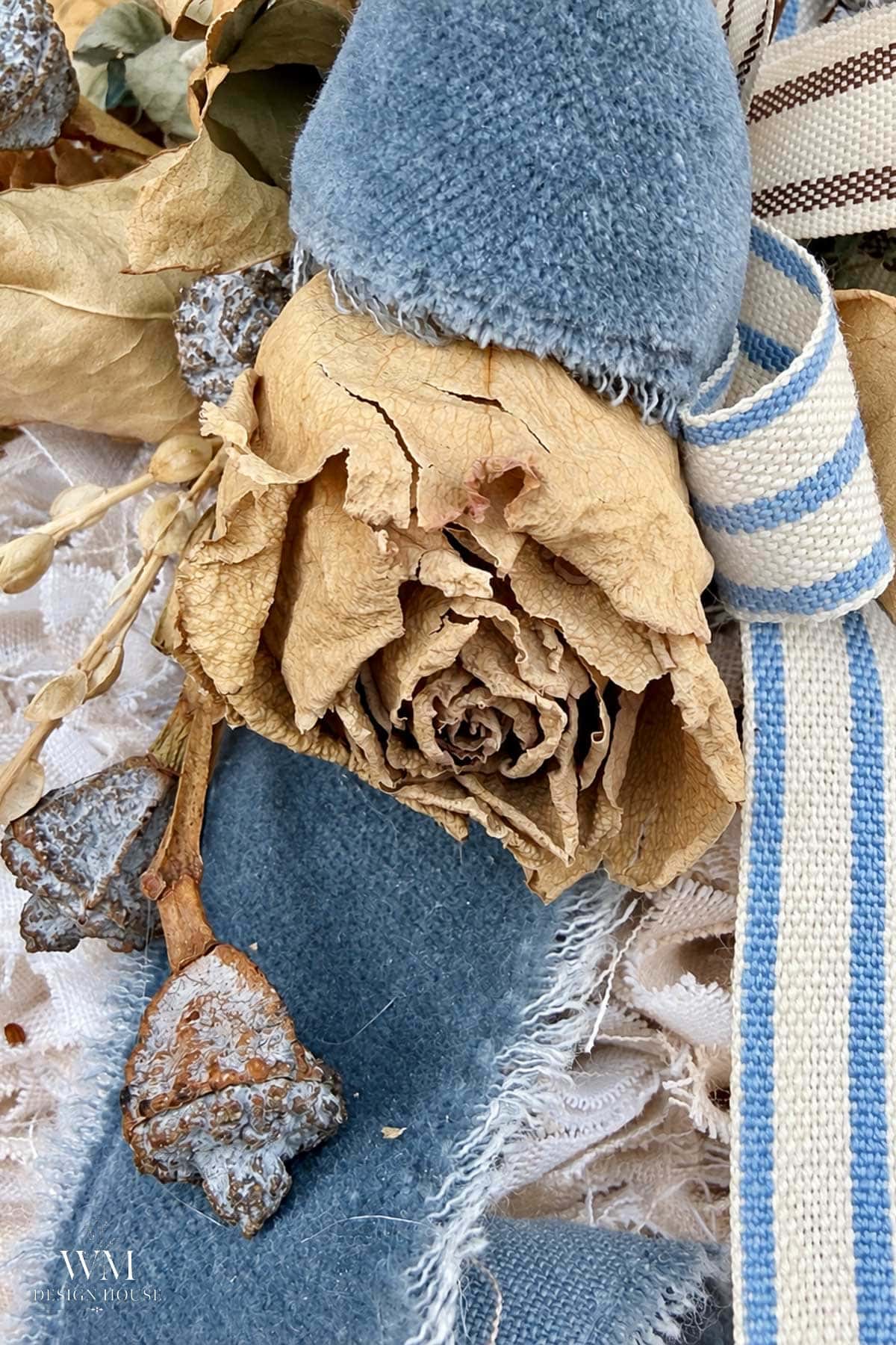 A dried beige rose surrounded by blue velvet fabric, rustic seed pods, and blue-and-white striped ribbon that adorn the top of a no sew fabric pupkin.