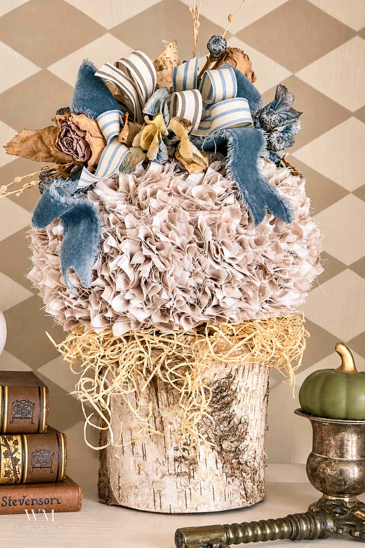 A no sew fabric pumpkin decoration with blue ribbon and dried flowers, sitting on a birch wood base next to stacked vintage books and a small green pumpkin.