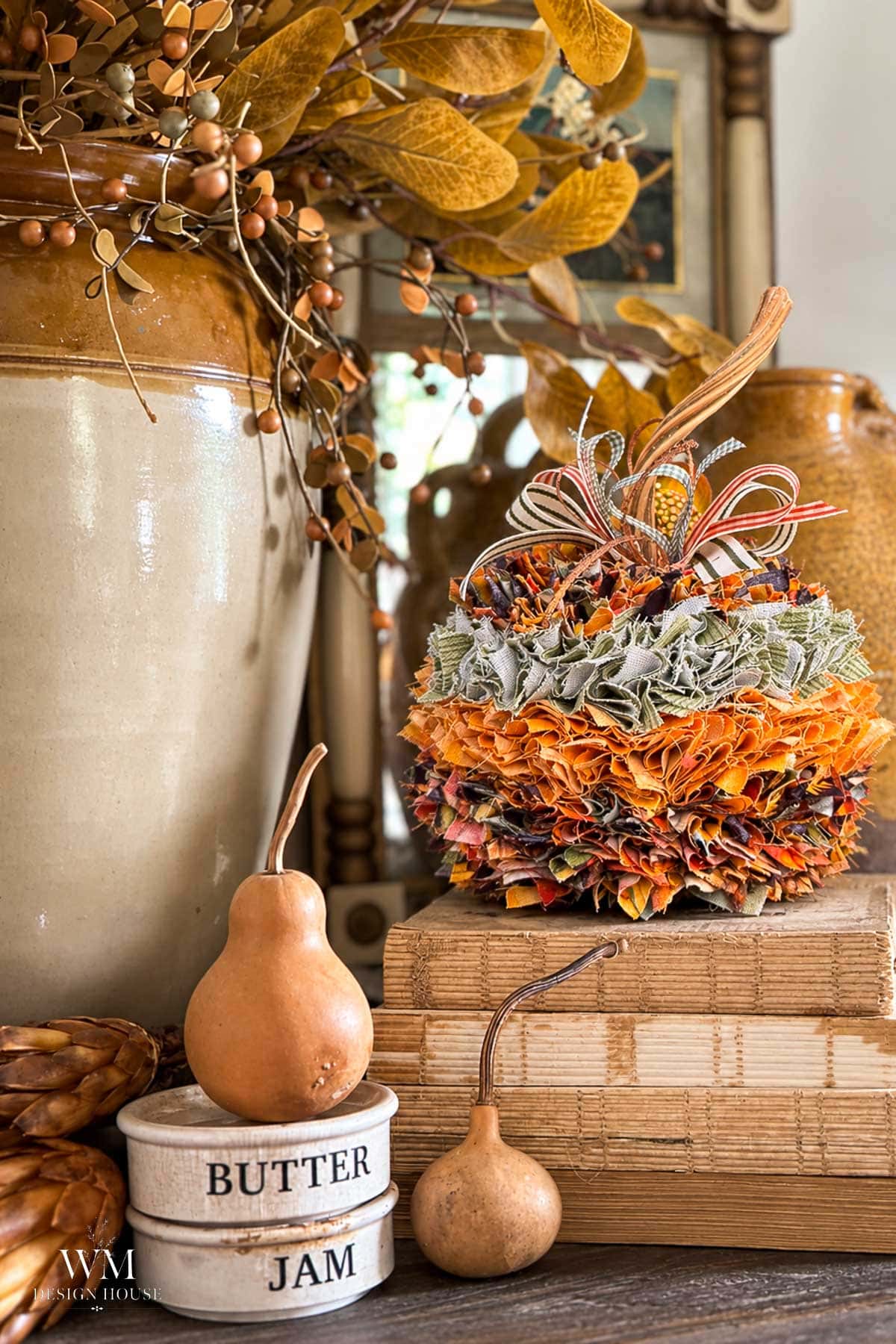 A decorative no sew fabric pumpkin with a ribbon bow, sitting on a stack of books next to a small gourd and a “Butter Jam” jar.