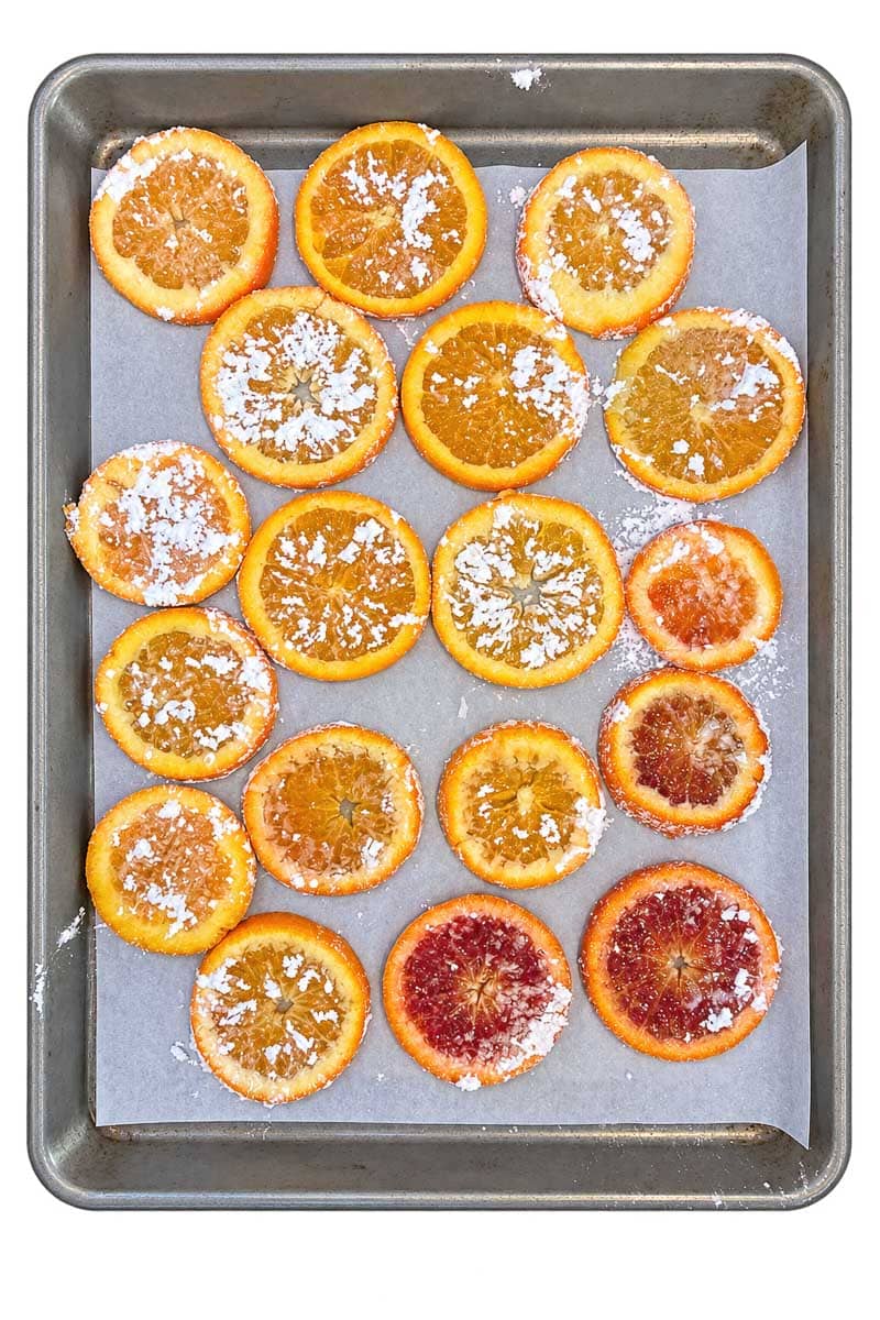 A baking sheet lined with parchment paper holds several dehydrated orange slices, some sprinkled with powdered sugar.