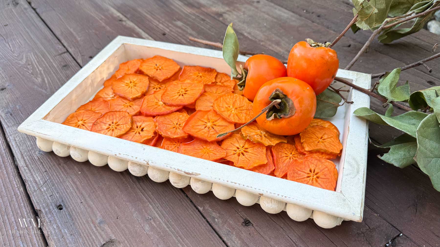 How to dry persimmons: a white tray of dried persimmon slices with two whole persimmons on the vine.