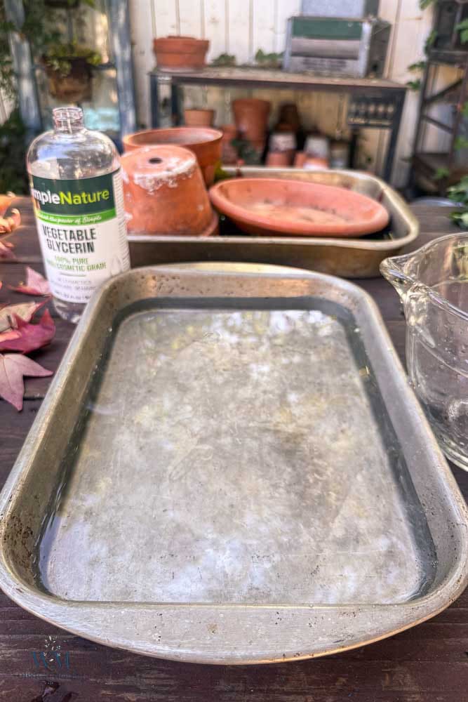 A rectangular metal baking pan filled with water, positioned next to a bottle of vegetable glycerin on a wooden surface.