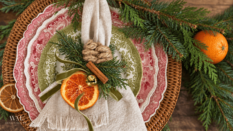 A place setting featuring patterned red and green plates on a woven charger, topped with a linen napkin held by a knotted rope napkin ring, accented with a dried orange slice, a cinnamon stick, and a jingle bell.