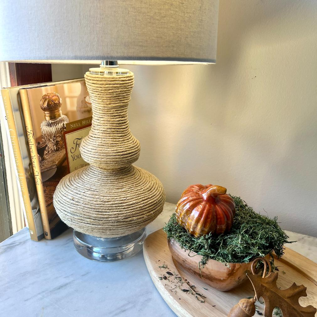 A rope-wrapped table lamp sits next to two books and a decorative ceramic pumpkin nestled in greenery on a wooden tray.