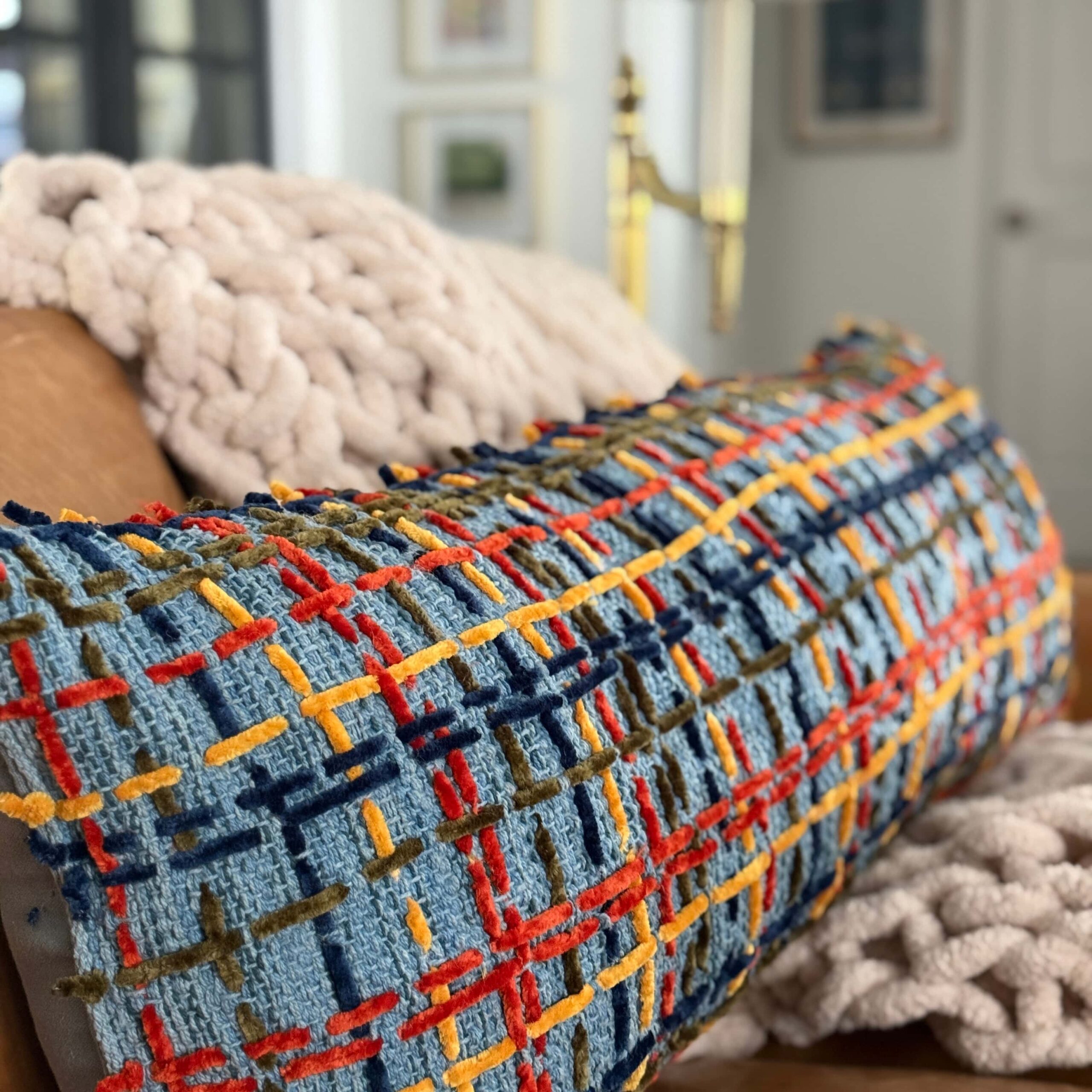 A rectangular decorative pillow with a blue textured grid pattern in red, yellow, and dark green, resting on a leather sofa with chunky knit throws.