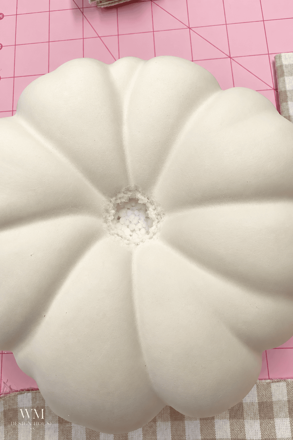 Top-down view of a cream-colored foam pumpkin for a no sew fabric pumpkin project, sitting on a pink cutting mat with a small hole in the center showing the white styrofoam interior with no stem.