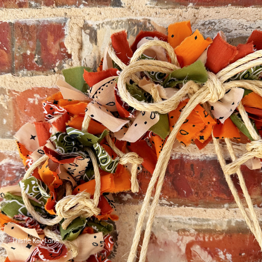 A rag wreath made of orange, green, and cream bandana scarves tied with jute rope, hanging against a brick wall.